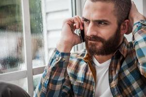 Bad news. Depressed young bearded man expressing negativity while talking on the mobile phone and looking through window photo