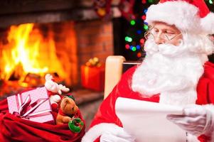todos recibirán un regalo. santa claus tradicional mirando su saco con regalos y sosteniendo un papel mientras se sienta en su silla con chimenea y árbol de navidad en el fondo foto