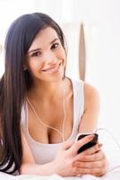 Enjoying her favorite music. Beautiful young smiling woman listening to the MP3 player and looking at camera while lying in bed photo