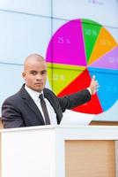 Look at the diagram Confident African man in formalwear standing at the tribune and pointing projection screen while making a presentation photo