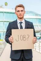 I need a job Handsome young man in formalwear holding poster with job text message while standing outdoors and against building structure photo