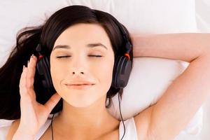 Woman enjoying music. Top view of beautiful young smiling woman adjusting her headphones while listening to the MP3 player in bed photo