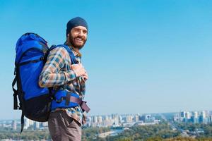 In a search of adventures. Handsome young man carrying backpack and looking at camera through the shoulders a with smile photo