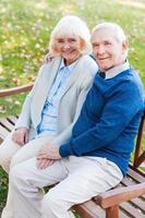 disfrutando el uno del otro. vista superior de la feliz pareja de ancianos tomándose de la mano y mirando la cámara con una sonrisa mientras se sientan juntos en el banco del parque foto