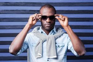 Perfect style. Handsome young African man adjusting his sunglasses while standing against striped background photo