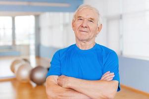 Confident and healthy. Confident senior man keeping arms crossed and looking at camera while standing in health club with sports equipment laying in the background photo