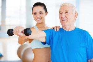 You are doing well Cheerful female physical therapist helping senior man with fitness in health club photo