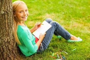 Little artist. Rear view of cute little blond hair girl drawing something at her note pad and looking over shoulder with smile while sitting on green grass and leaning at the tree photo