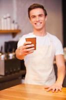 sirviendo café para llevar. apuesto joven barista estirando la mano con una taza de café y sonriendo mientras se inclina en el mostrador del bar foto