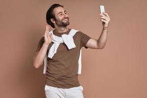 joven feliz mirando su teléfono inteligente y agitando la mano contra el fondo marrón foto