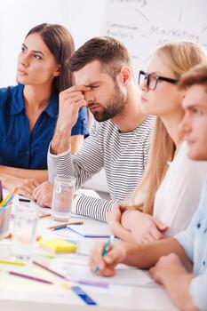 Tired and stressed. Depressed young man touching face with hand while sitting at the table together with his colleagues photo