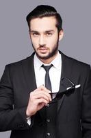 Confident and good looking. Portrait of handsome young man in formalwear holding sunglasses and looking at camera while standing against grey background photo