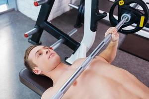 Training with weights. Top view of young muscular man working out on bench press photo