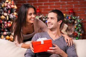 Christmas is time for giving. Beautiful young woman giving a red gift box to her boyfriend and smiling with Christmas Tree in the background photo