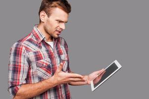 It can not be truth Disappointed young man holding a digital tablet and pointing it after reading bad news, while standing against grey background photo