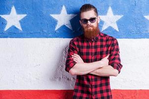 American style. Handsome young bearded man in sunglasses keeping arms crossed and looking at camera while standing against American flag photo