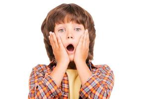 Surprised little boy. Shocked little boy keeping mouth open and touching face with hands while standing isolated on white photo