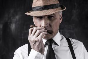My favorite cigar. Confident senior man in hat and suspenders smelling cigar while standing against dark background photo