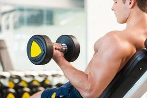 Training with dumbbells. Rear view of confident young muscular man training with dumbbell in gym photo