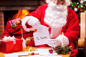 Papá Noel en el trabajo. primer plano de santa claus leyendo cartas mientras está sentado en su silla con un árbol de navidad y una chimenea en el fondo foto