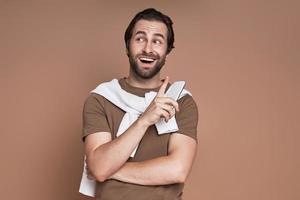 Excited young man holding smart phone while standing against brown background photo