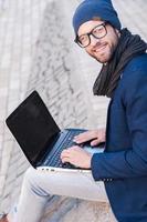 Surfing the net outdoors. Top view of handsome young man in smart casual wear working on laptop and smiling while sitting outdoors photo