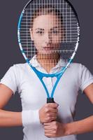 I know how to win Beautiful young women in sports clothes holding tennis racket in front of her face and looking at camera while standing against grey background photo