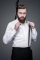 Unforeseen fail. Frustrated young bearded man adjusting his suspenders and keeping eyes closed while standing against grey background photo