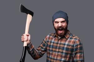 Furious lumberjack. Furious young bearded man swinging his axe and shouting while standing against grey background photo