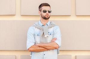 Cool and handsome. Handsome young man in sunglasses keeping arms crossed and looking away while standing in front of the textured wall outdoors photo