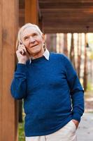 Estoy muy feliz de saber de usted, un anciano feliz hablando por teléfono móvil y sonriendo mientras está parado al aire libre. foto