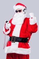 Listening to the Christmas music. Santa Claus in sunglasses and headphones listening to MP3 Player and gesturing while standing against grey background photo