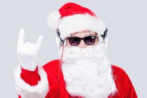 Papá Noel genial. santa claus con gafas de sol y auriculares escuchando un reproductor de mp3 y gesticulando mientras se enfrenta a un fondo gris foto