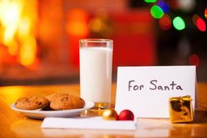 para papá noel primer plano de galletas y vaso de leche en la mesa con árbol de navidad y chimenea en el fondo foto