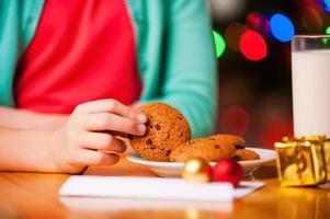 mis galletas navideñas favoritas primer plano de una niña tomando una galleta del plato mientras está sentada en la mesa con un árbol de navidad en el fondo foto