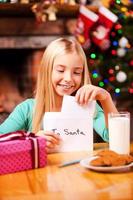 carta a papa noel linda niña poniendo una carta a santa en el sobre mientras se sienta en la mesa con un vaso de leche y galletas sobre ella foto