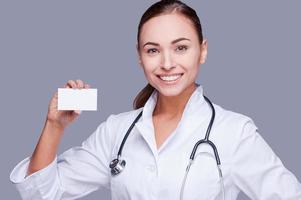 Copy space on her card. Confident female doctor in white uniform holding card and looking at camera and smiling while standing against grey background photo