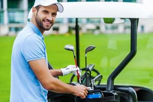 Choosing the proper driver. Handsome young male golfer choosing driver while standing near the golf cart photo