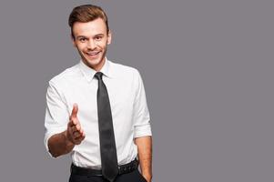 You are welcome in our company Portrait of cheerful young man in formalwear stretching out hand for shaking while standing against grey background photo