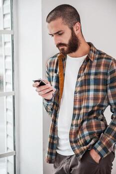 Typing text message. Confident young bearded man typing message on his smart phone while standing near the window photo