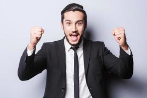 llegué al mejor joven feliz en ropa formal gesticulando y sonriendo mientras estaba de pie contra un fondo gris foto