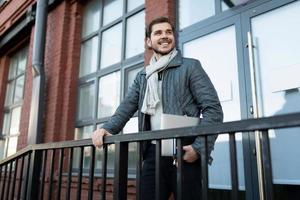 portrait of a cheerful man in autumn clothes with a laptop against the background of a brick building with large windows photo