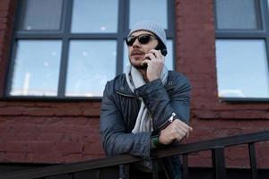 a man in a hat and sunglasses speaks on a mobile phone in front of a brick building with large windows photo
