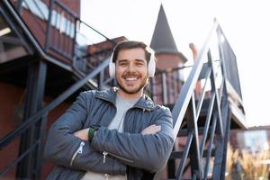 an adult man with a broad smile on his face listens to music in headphones with his arms crossed on his chest against the backdrop of stairs outside the office photo