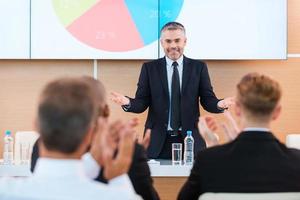 Celebrating success. Confident mature man in formalwear gesturing and smiling while making presentation in conference hall with people applauding on the foreground photo