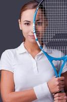 I am in game. Beautiful young women in sports clothes holding tennis racket in front of half of her face and looking at camera while standing against grey background photo