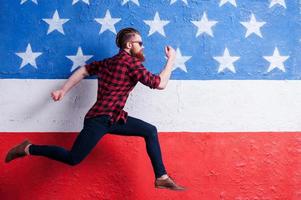 Hurrying to be in trend. Handsome young bearded man wearing sunglasses and running along American flag photo