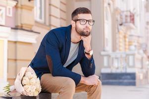 esperando a su novia. un joven pensativo con una chaqueta inteligente sosteniendo la mano en la barbilla mientras se sienta en el banco con un ramo de flores cerca de él foto