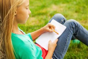 Performing her creativity. Cute little blond hair girl drawing something at her note pad while sitting on green grass and leaning at the tree photo