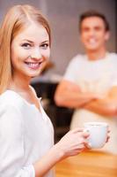 Happy customer. Beautiful young woman holding cup of coffee and smiling while standing near bar counter and with cheerful barista standing in the background photo